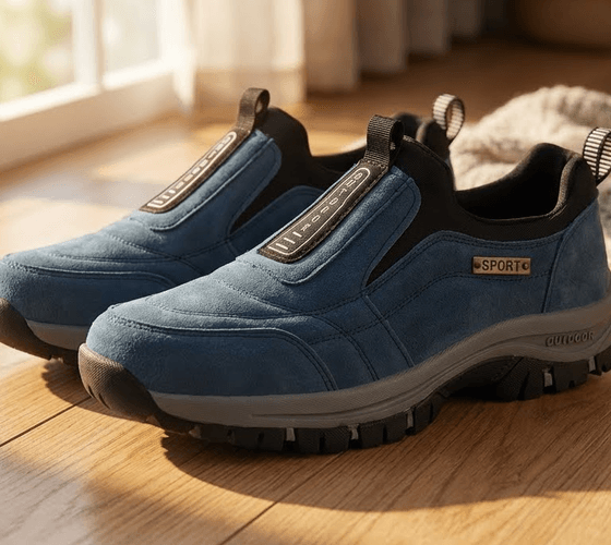 A pair of blue Stepprs slip-on shoes sitting on a wooden floor in warm indoor light beside a folded throw blanket.